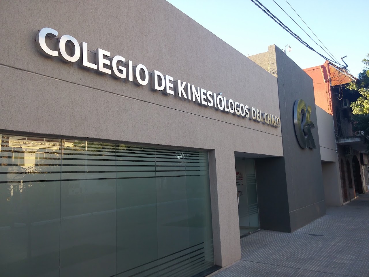 Modern institutional architectural exterior building facade of a professional medical or regulatory college headquarters in Resistencia Argentina, elegant clean stone and glass entrance, subtle institutional lettering, warm sunlight, manicured entrance greenery, high-end professional architectural photography, crisp and welcoming.